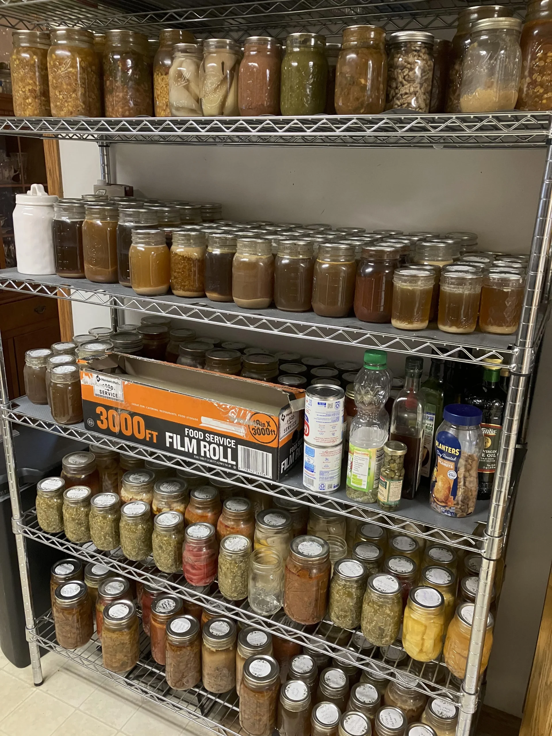 A metal shelving unit holds rows of glass jars filled with preserved foods and liquids, a box of film roll, canned goods, bottles, and other kitchen items organized neatly on multiple shelves.