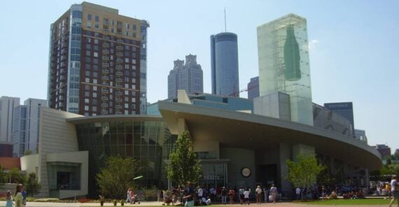 People gather on a grassy lawn in front of a modern, glass-fronted building with a curved roof and a tall glass structure, against a backdrop of tall city skyscrapers under a clear sky.