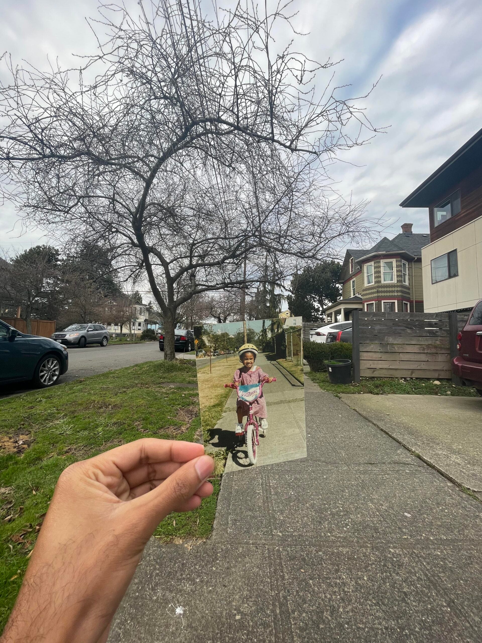A hand holds a photo of a child riding a pink bike, aligning it with the same sidewalk and tree in the present-day street scene, blending past and present. Leafless trees and houses appear in the background.
