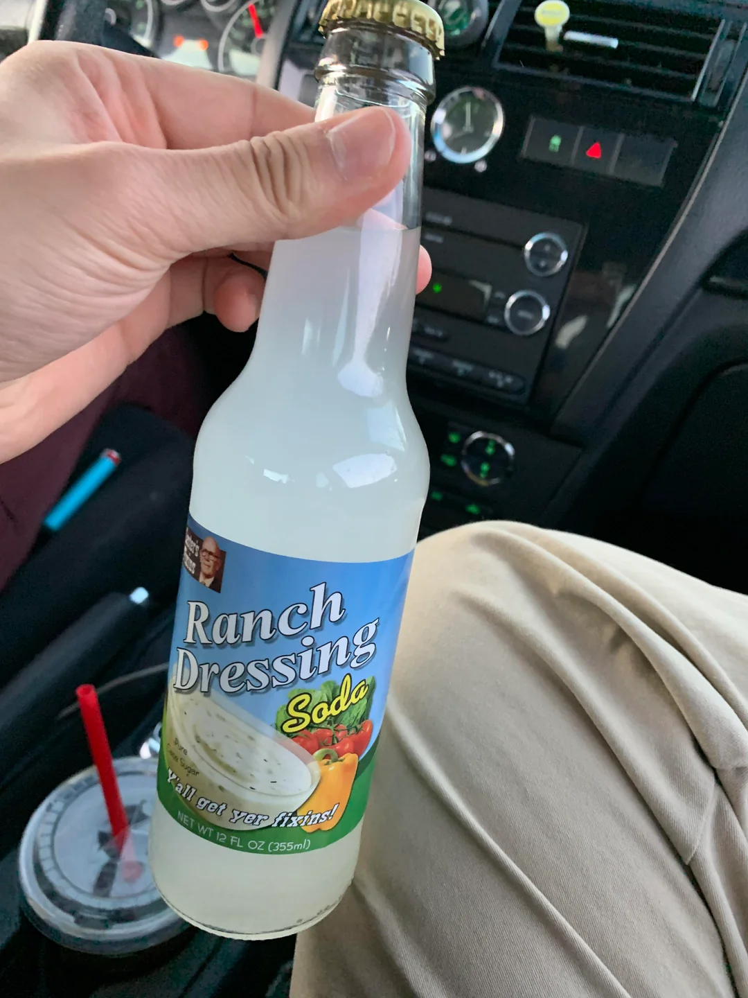 A hand is holding a clear glass bottle labeled "Ranch Dressing Soda" with a picture of ranch dressing and vegetables on the label. The bottle contains a cloudy, white liquid. A car interior is visible in the background.