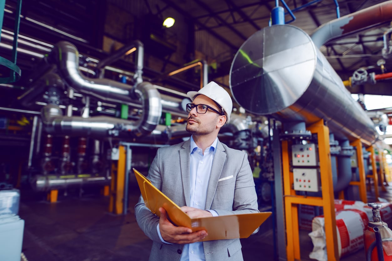A man in a suit and white hard hat holds a folder and inspects equipment inside an industrial facility with large pipes and machinery.