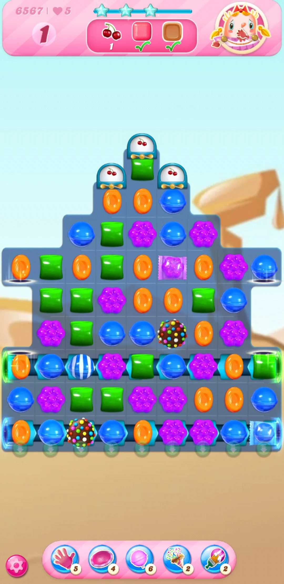 A colorful Candy Crush game board with various colored candies, special candies, and blockers arranged in a pyramid shape. The top shows two cherries to collect and five lives remaining.