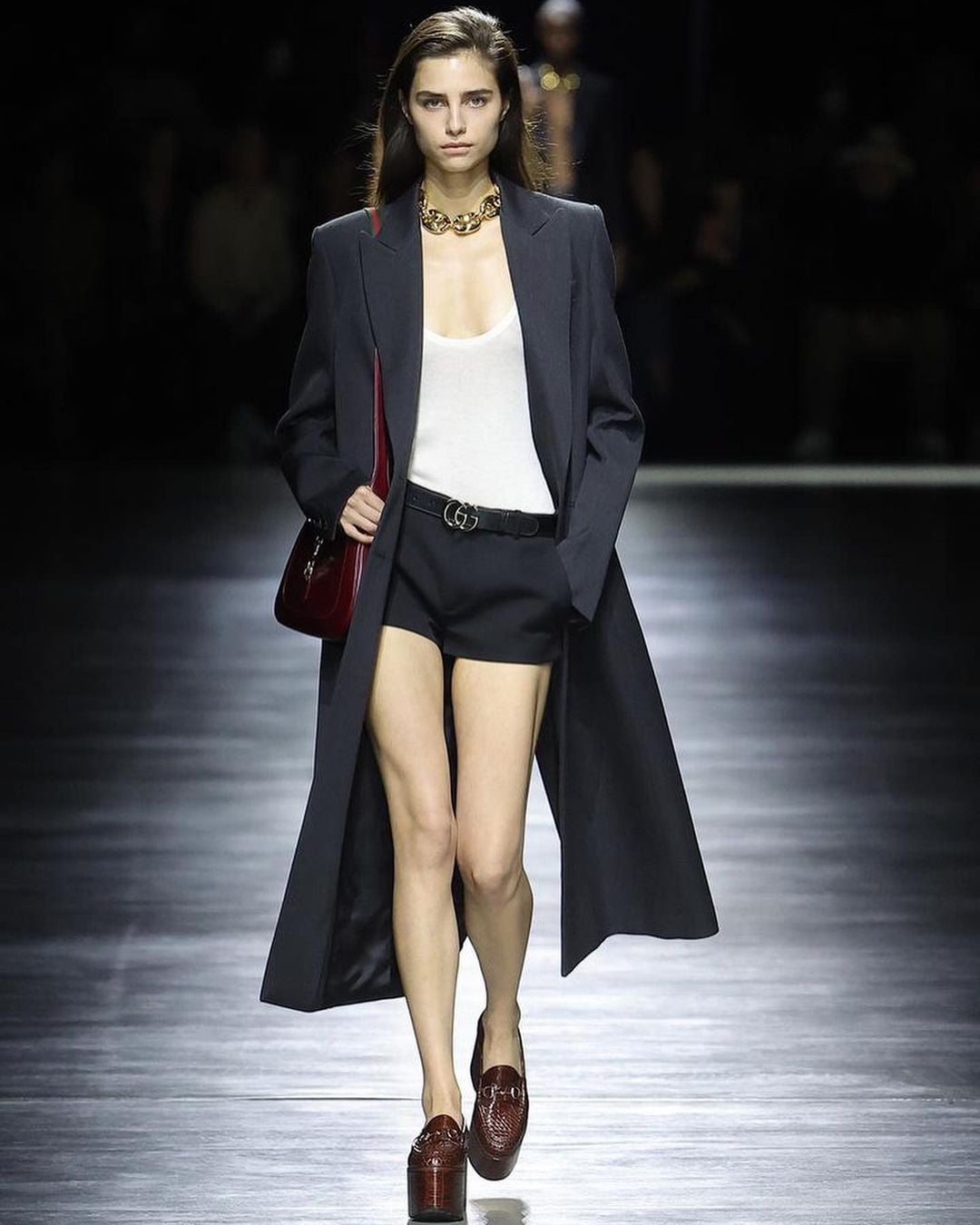 A model walks a runway wearing a long black coat, white tank top, black shorts with a belt, brown platform loafers, a chunky gold necklace, and carrying a dark red shoulder bag. The background is dark and blurred.