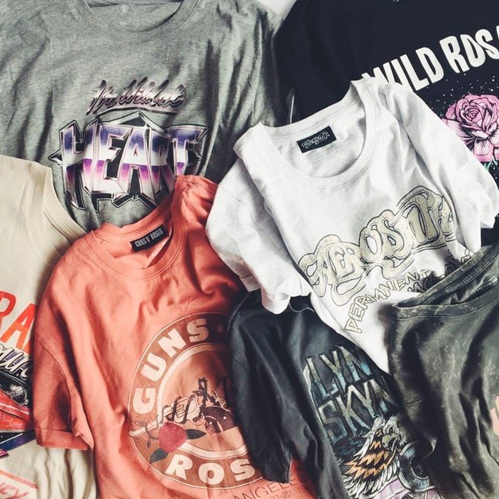 A collection of colorful vintage band t-shirts, including designs for Heart, Guns N’ Roses, Aerosmith, and Lynyrd Skynyrd, laid out overlapping on a flat surface.