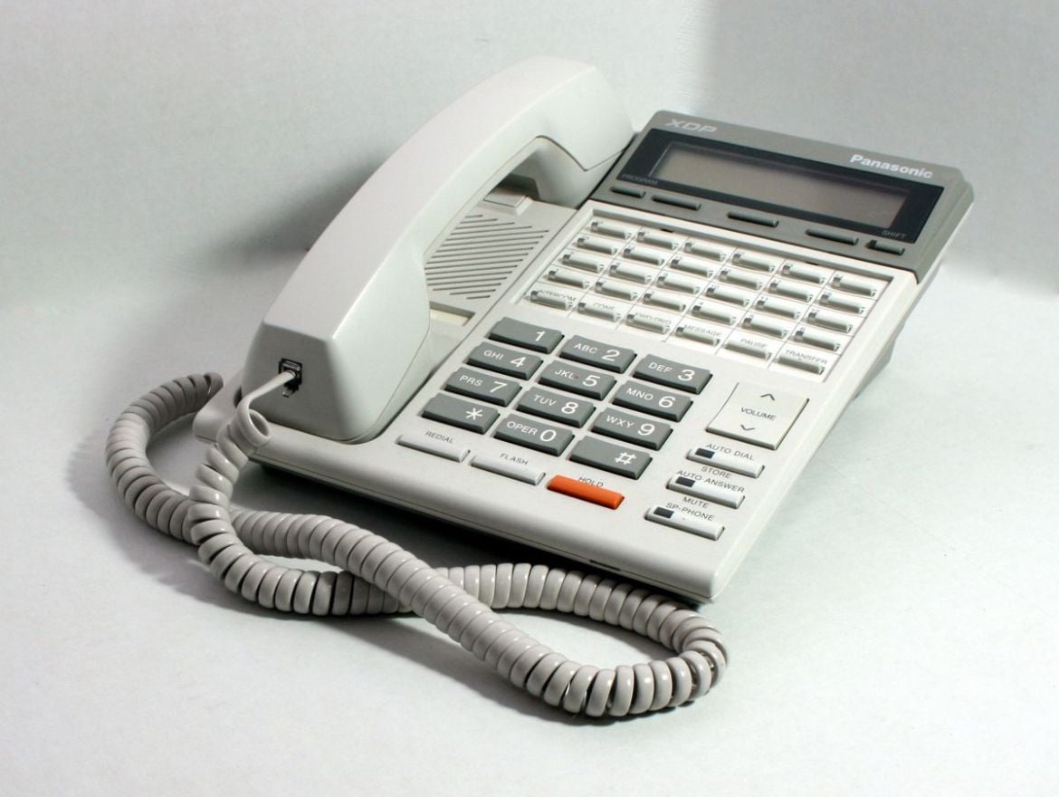A white corded telephone with a numeric keypad, function buttons, and a coiled handset cord resting on a light surface. The phone is labeled "Panasonic" and features a small display screen.