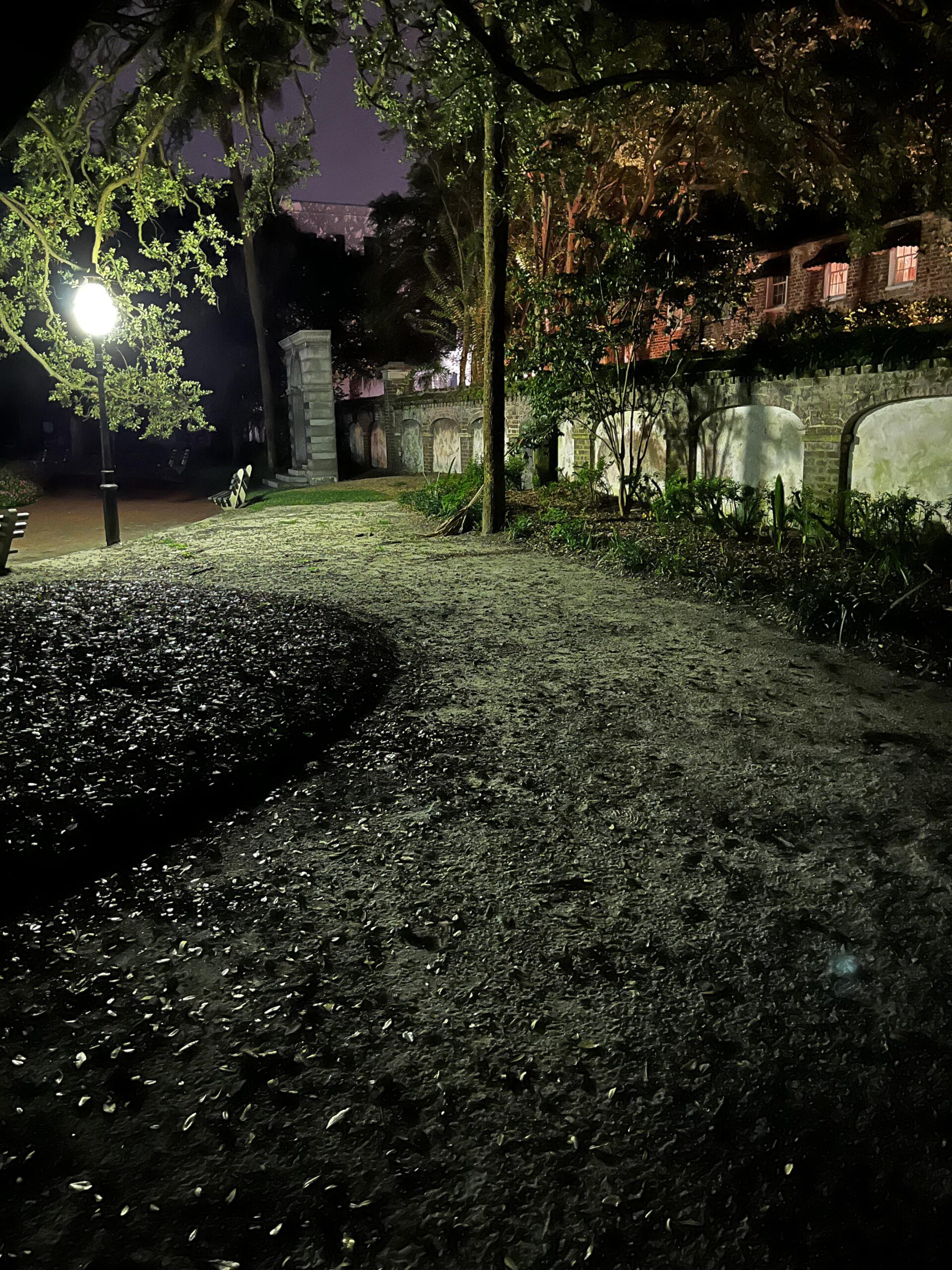 A dimly lit park at night with a glowing streetlamp, a dirt path, trees, benches, and an old stone wall. The scene has a moody, quiet atmosphere, with light spilling onto the walkway and shadows among the greenery.
