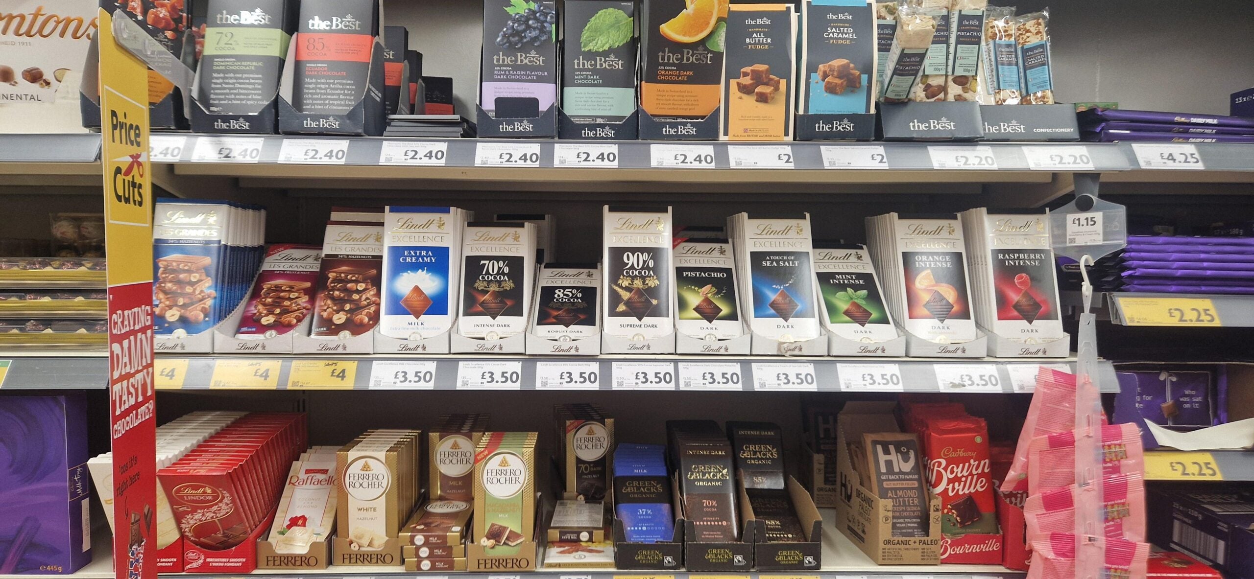 Shelves in a store display various brands of chocolate bars, including Lindt, The Best, and others. The chocolates are arranged in rows with visible price labels beneath each product.