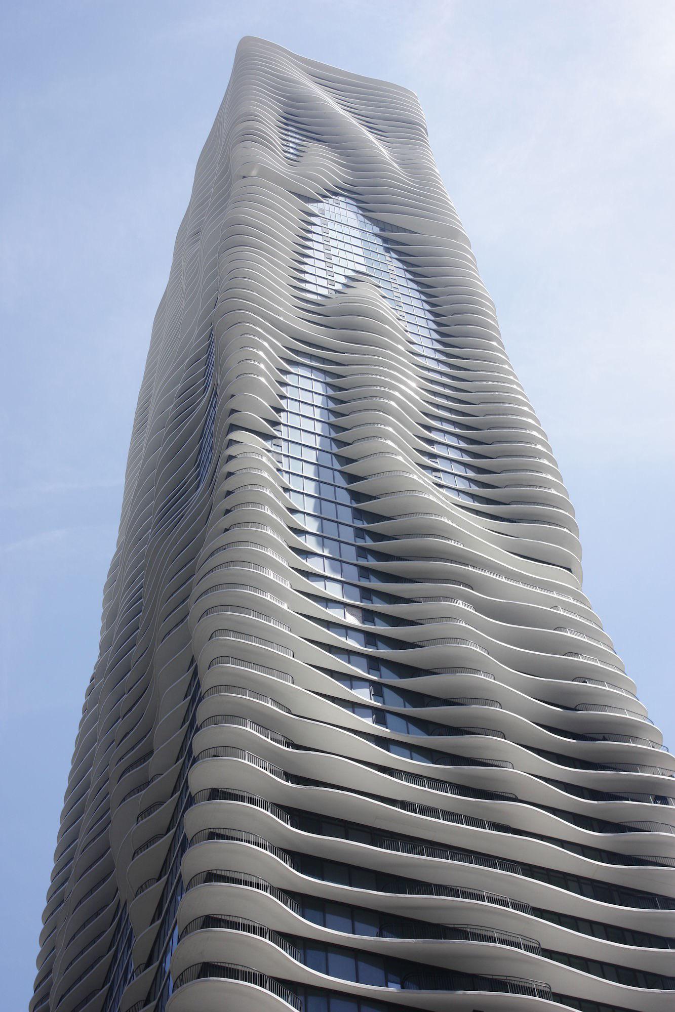 A tall modern skyscraper with undulating, wave-like balconies and a glass facade, viewed from below against a pale blue sky.