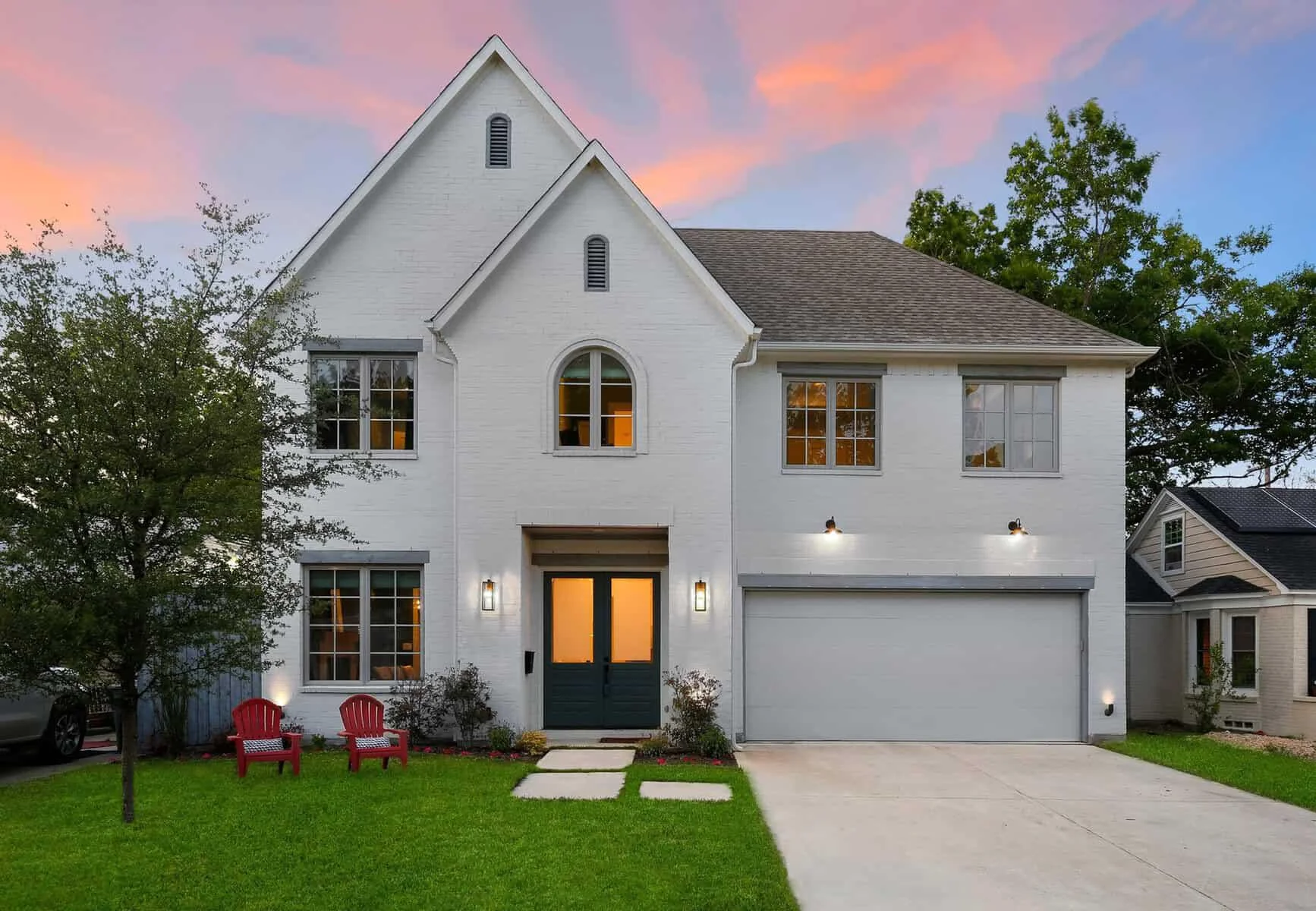 A modern two-story white brick house with large windows, a double garage, and two red Adirondack chairs on a green lawn at sunset. Trees surround the yard, and the sky is pink and orange.