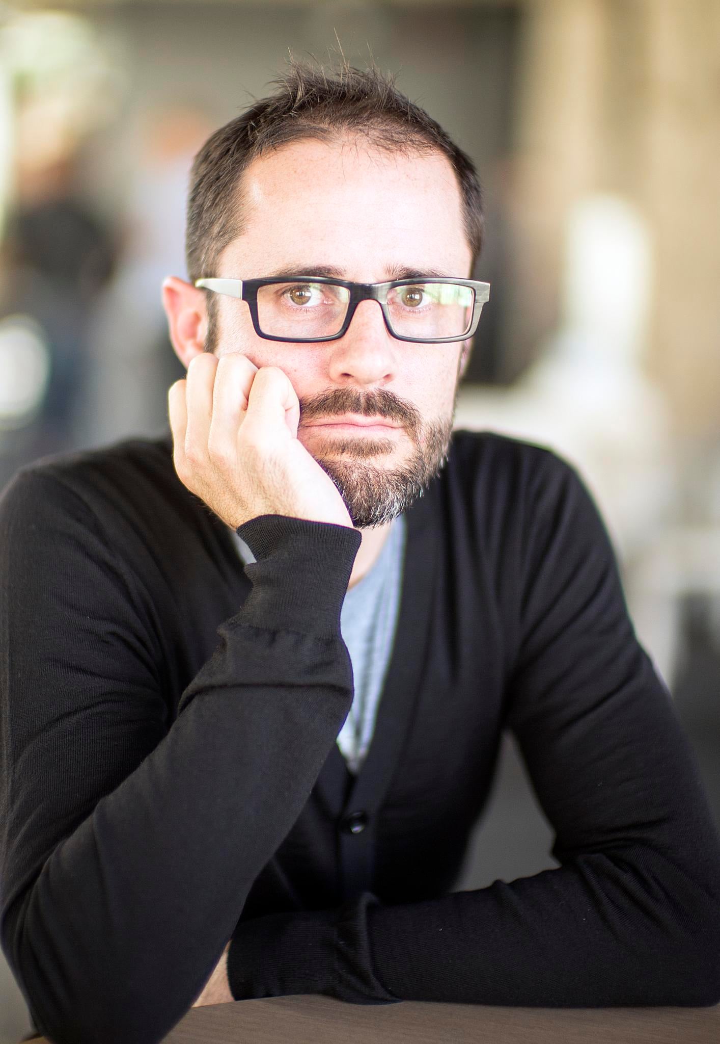 A man with short dark hair, a beard, and glasses rests his chin on his hand, wearing a black sweater, looking thoughtfully at the camera. The background is softly blurred.