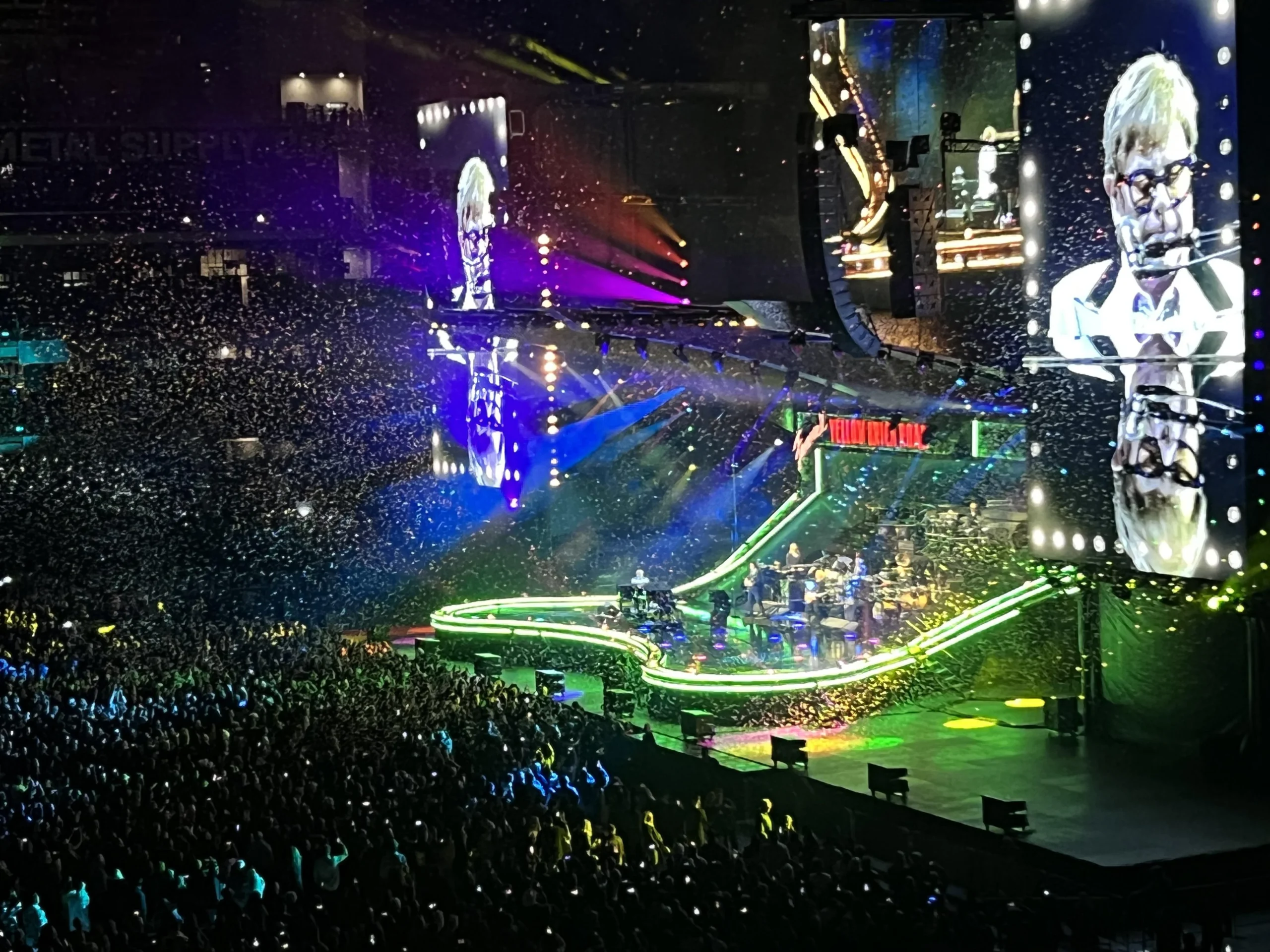 A large crowd watches a colorful concert in a stadium, with bright lights and confetti filling the air. A performer is visible on stage and on a big screen, playing piano in a white and black outfit.