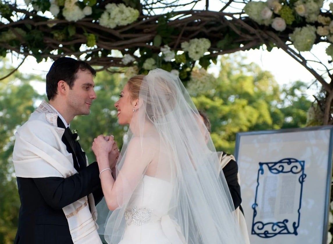 A bride and groom stand under a floral wedding canopy, holding hands and smiling at each other. The bride wears a white dress and veil, and the groom wears a suit with a white shawl. A framed document is visible beside them.