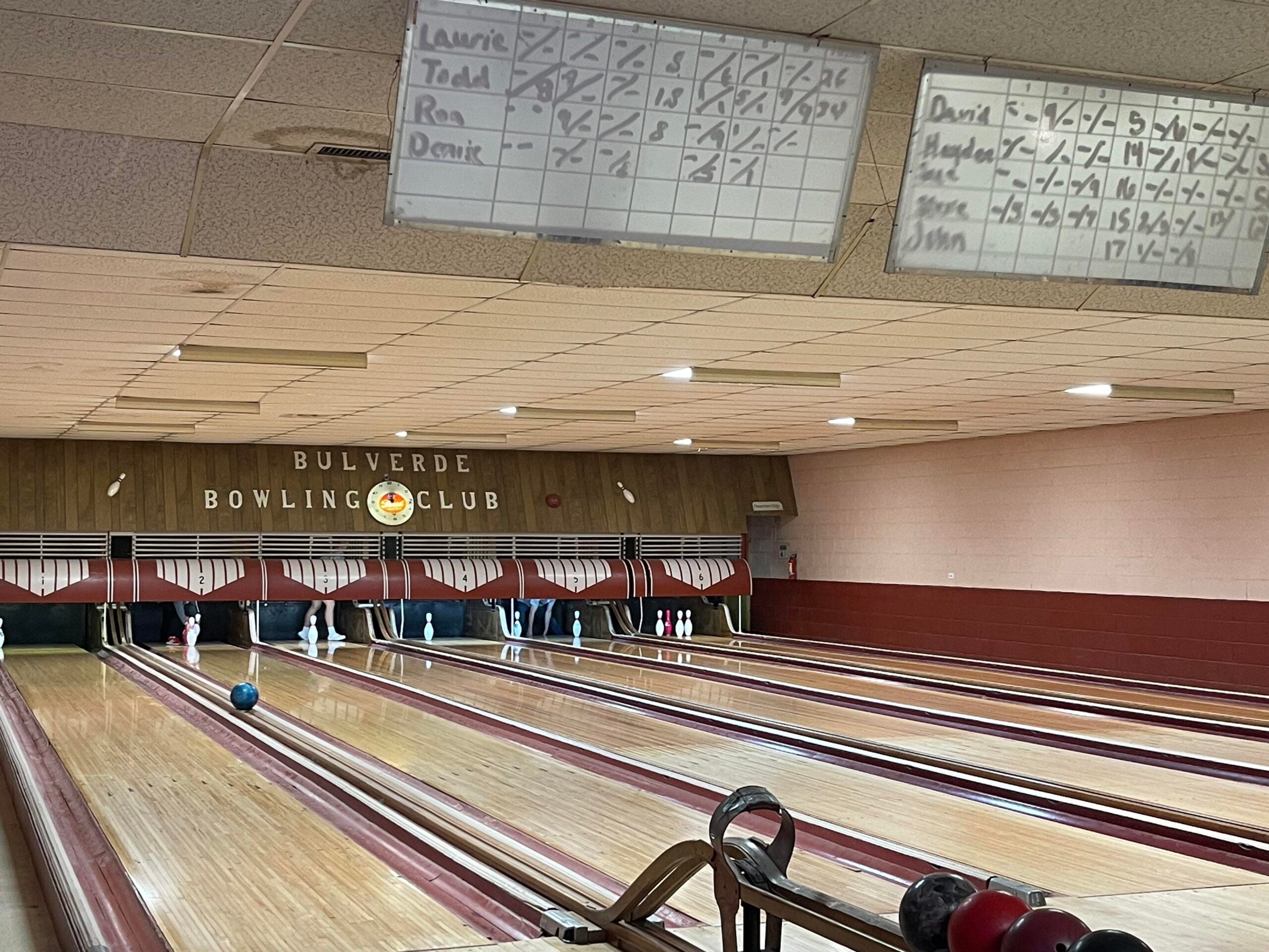 Several bowling lanes inside Bulverde Bowling Club, with pins set up and a blue bowling ball rolling down one lane. Two scoreboards with handwritten scores are visible above the lanes.