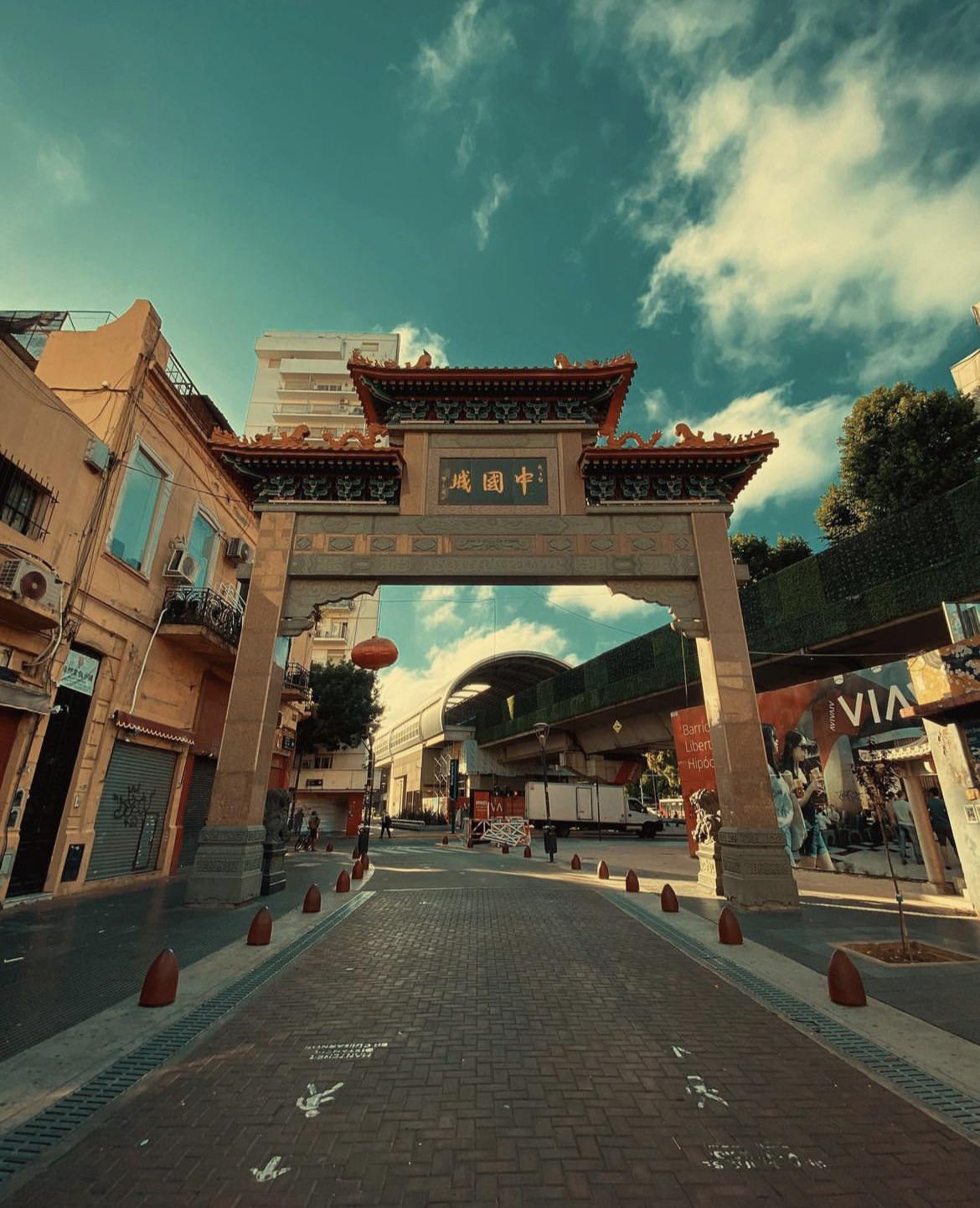 A traditional Chinese archway stands at the entrance of a street lined with buildings under a blue sky with scattered clouds. Red lanterns hang from the arch, and the street is quiet with no visible people.