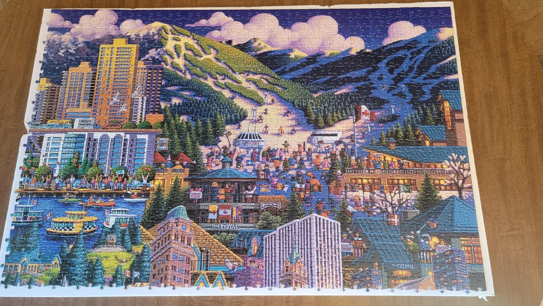 A colorful completed jigsaw puzzle shows a lively mountain town with tall buildings, boats on a lake, snowy mountains, evergreen trees, and crowds of people enjoying various winter and urban activities.