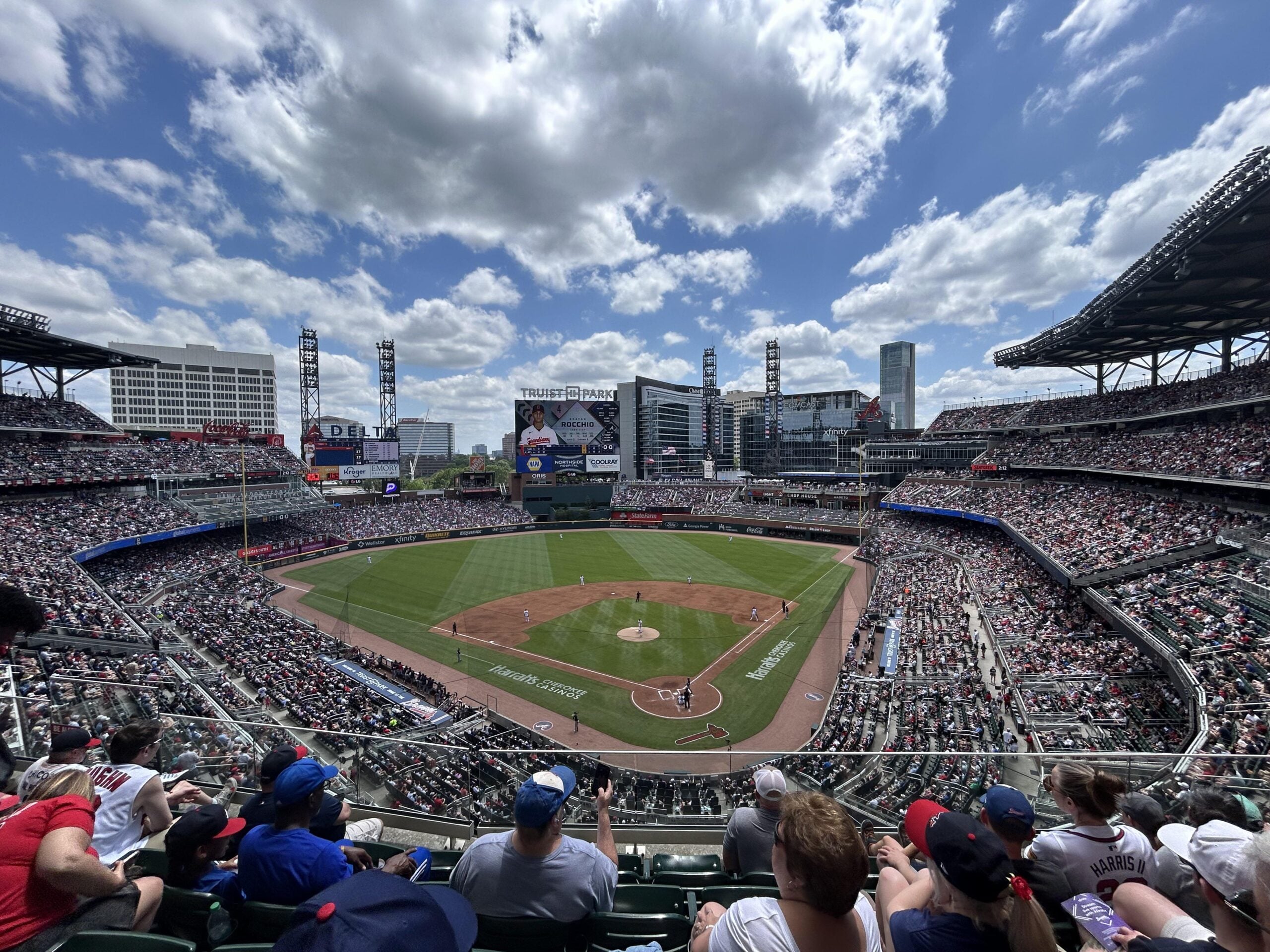 Wide-angle view of a packed baseball stadium under a partly cloudy sky, showing the field, players, and crowds in the stands. The scoreboard and city buildings are visible in the background.