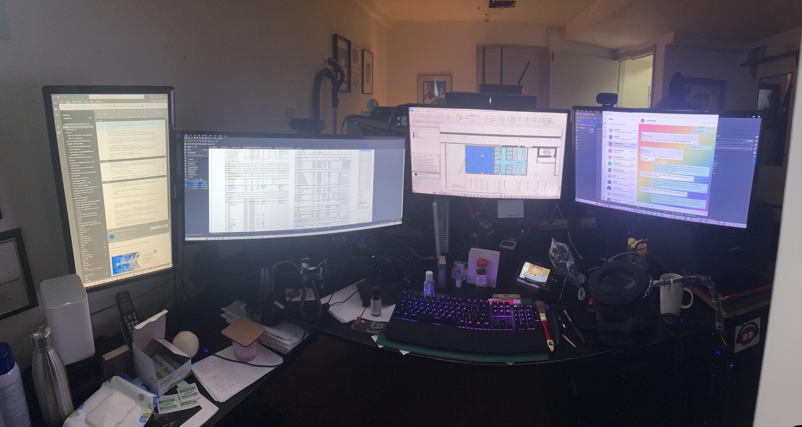A wide computer desk with four monitors displaying spreadsheets, documents, and messaging apps. The desk is cluttered with a keyboard, mouse, speakers, papers, bottles, and various office items.