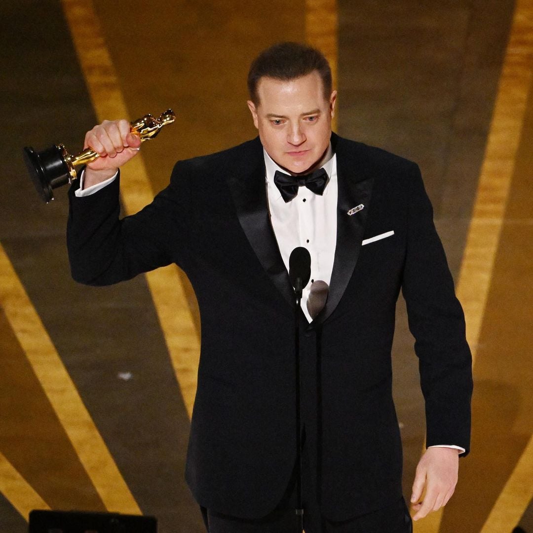 A man in a black tuxedo holds up an Oscar trophy while standing on stage, speaking into a microphone, with golden lines radiating on the floor behind him.