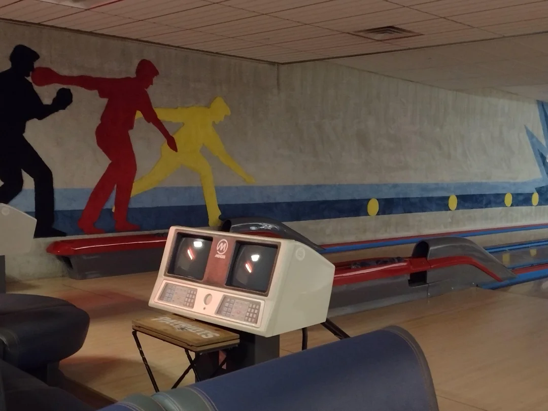 A vintage bowling alley with old-style scoring monitors and a mural on the wall showing red, black, and yellow silhouettes of people bowling.