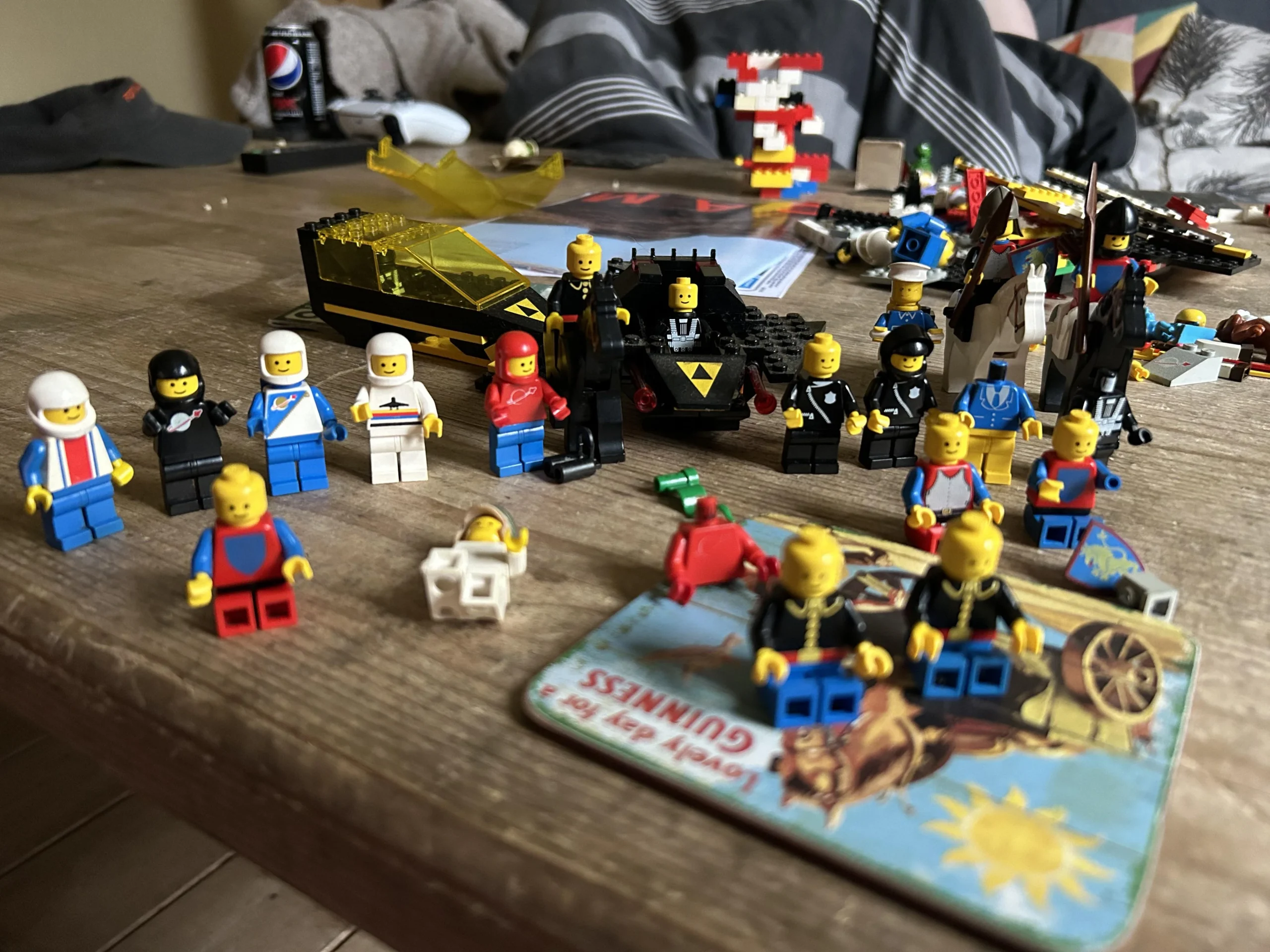 A group of LEGO minifigures are arranged on a wooden table with various LEGO pieces, a coaster, and toys scattered around. A can of Pepsi and other items are visible in the background.