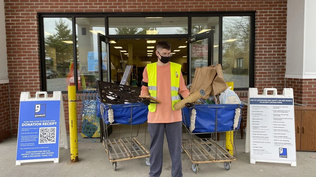 A person in a yellow safety vest and black mask stands outside a donation center, holding a grate and a brown bag. Two large blue carts of donations and donation instruction signs are visible beside the entrance.