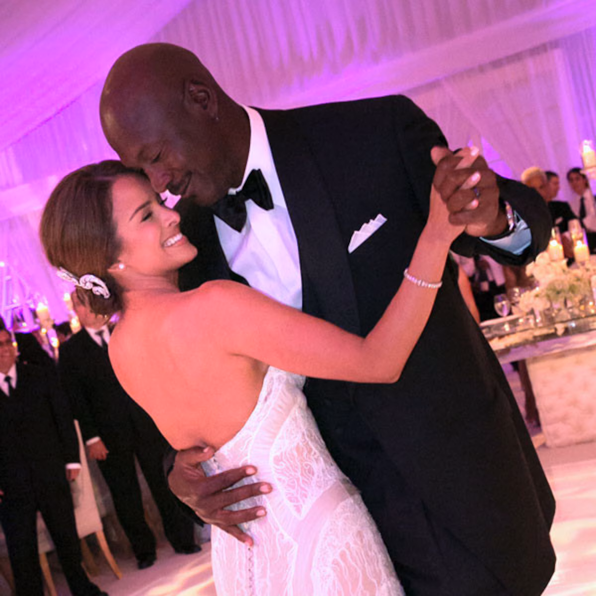 A bride in a strapless white gown and a groom in a black tuxedo smile and embrace while dancing at their wedding reception, surrounded by elegantly dressed guests and soft purple lighting.