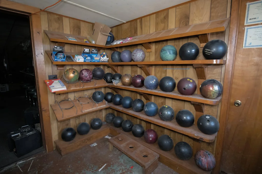 Wood-paneled room with shelves holding various bowling balls and some boxes. A wooden bench with foot holes is on the floor. Some certificates are visible on the wall. The room appears dimly lit and somewhat cluttered.
