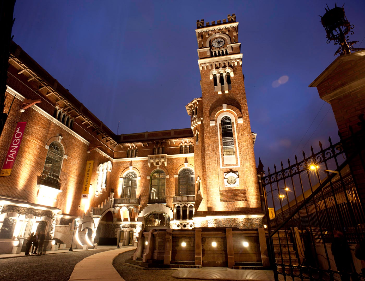 A historic brick building with arched windows and a tall clock tower is lit up at dusk. Several people stand near the entrance, and an ornate iron gate is visible in the foreground.