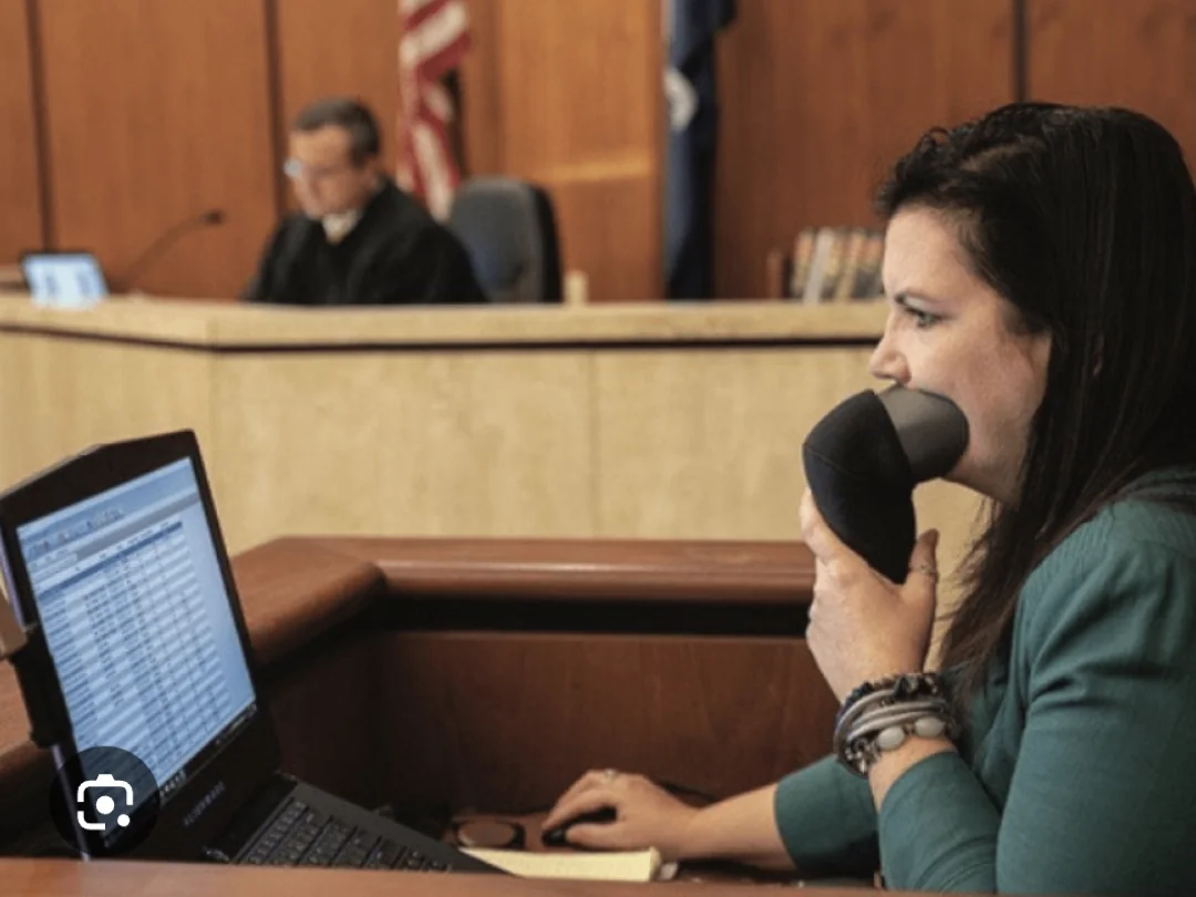 A court reporter sits at a desk typing on a laptop and speaking into a stenomask, while a judge is seated in the background on the courtroom bench.