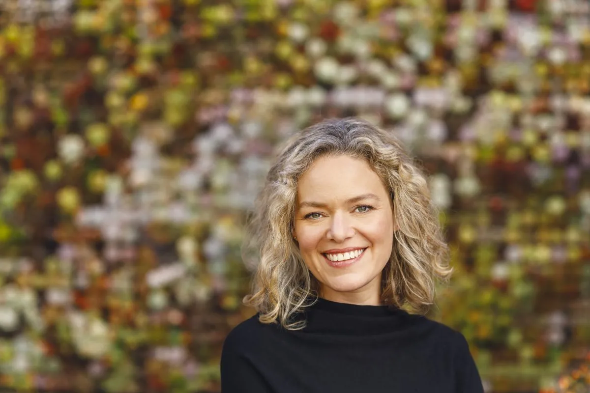 A woman with curly blonde hair wearing a black top smiles in front of a colorful, blurred floral background.