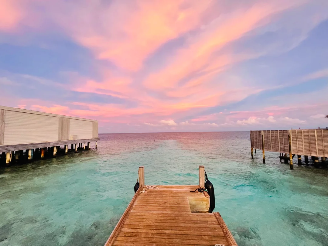 A wooden pier extends into clear turquoise water, flanked by overwater bungalows on both sides. The sky above glows with soft pink and orange clouds, suggesting a tranquil sunset or sunrise.