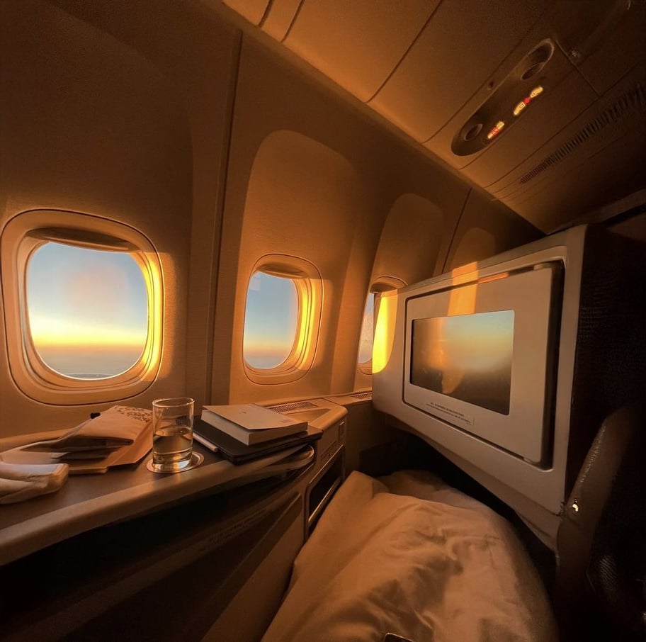 A cozy first-class airplane seat at sunset, with three windows showing golden light. A glass of water, books, and a napkin rest on the side table next to a screen and a neatly made bed.