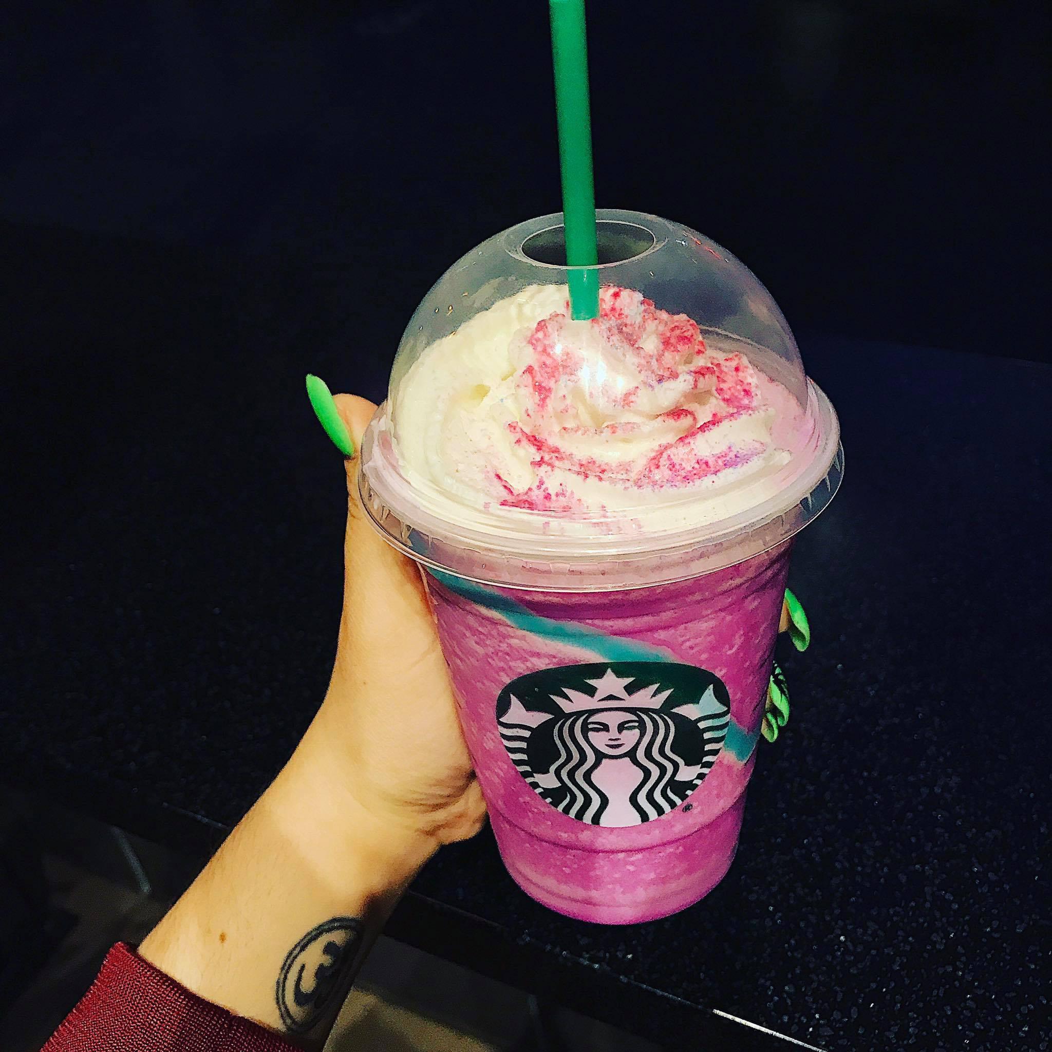 A hand holding a Starbucks cup filled with a bright pink blended drink, topped with whipped cream and pink sprinkles, with a green straw and a dome lid against a dark background.