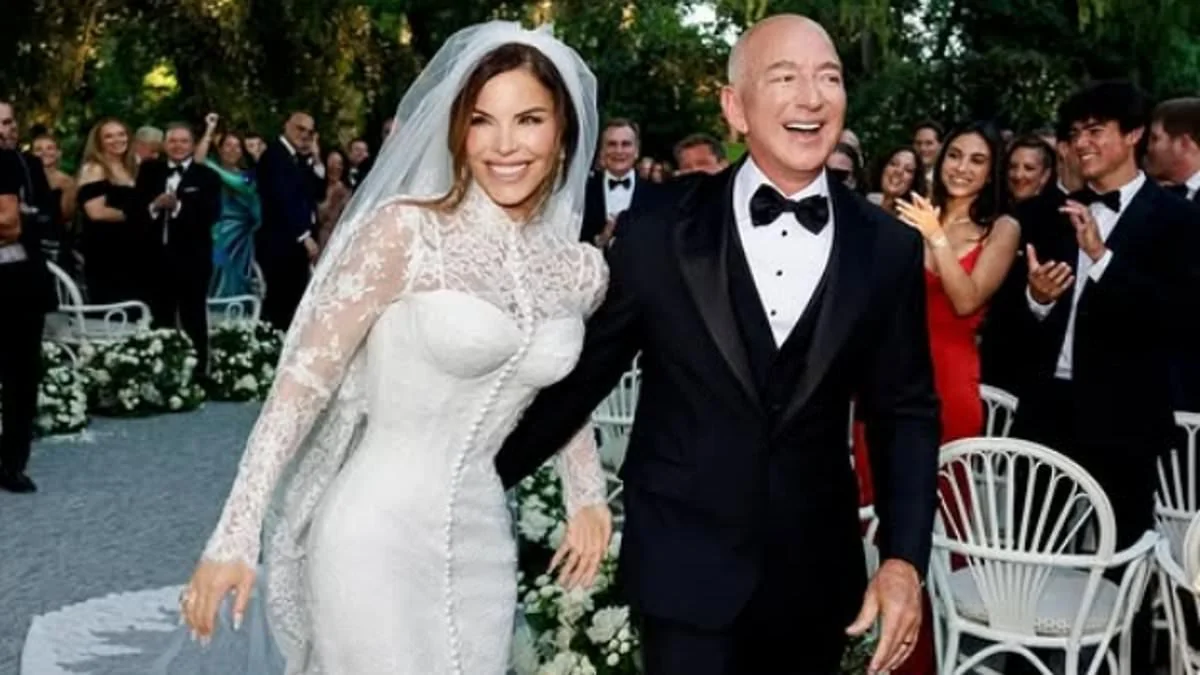 A bride in a white lace wedding dress and veil stands smiling beside a groom in a black tuxedo. They walk down the aisle outdoors, surrounded by applauding guests and white floral arrangements.