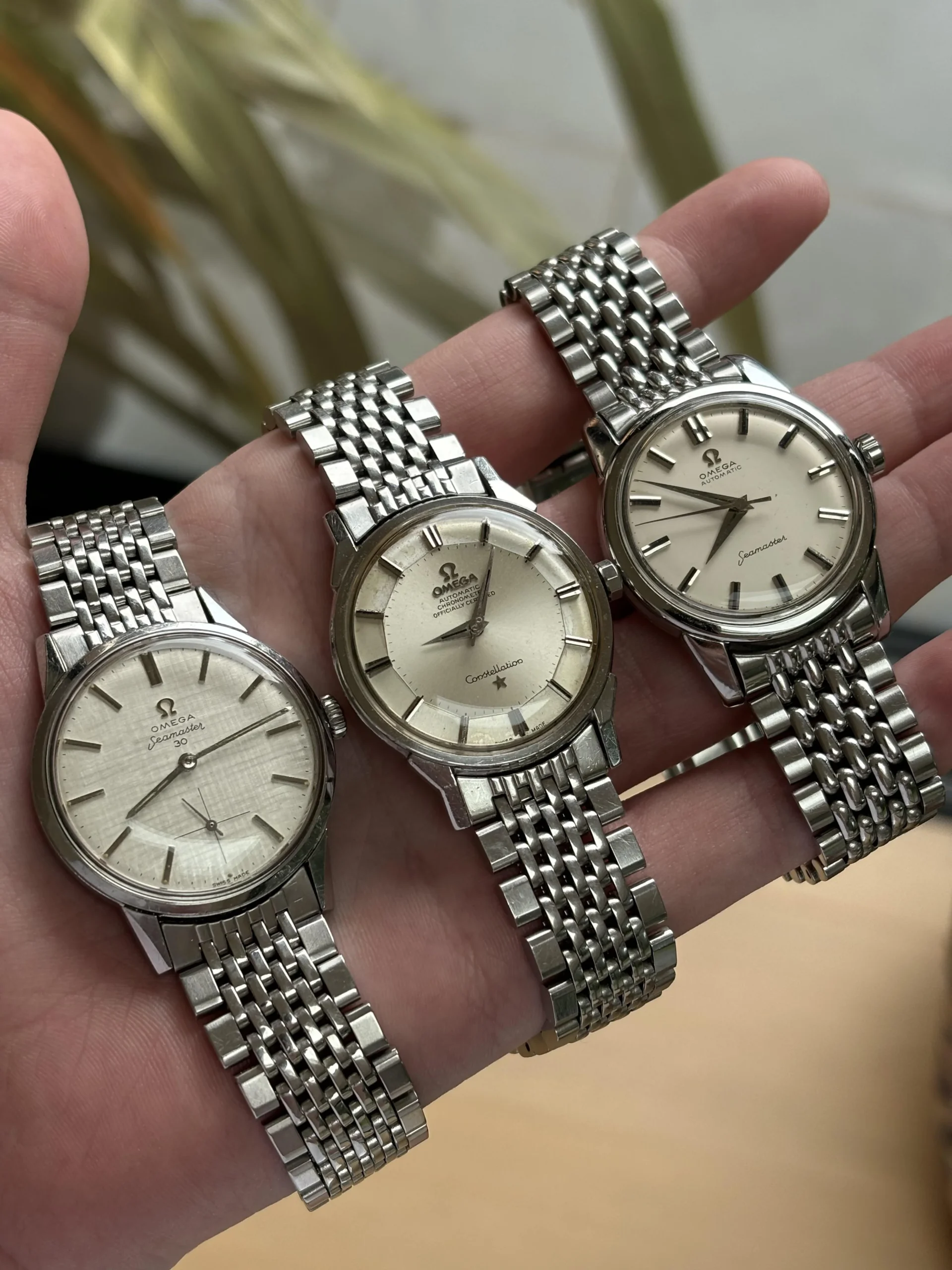 A hand holds three vintage Omega wristwatches with silver dials and metal link bracelets. The watches are displayed side by side, showing their classic designs and Omega logos. Plants and a blurred background are visible.