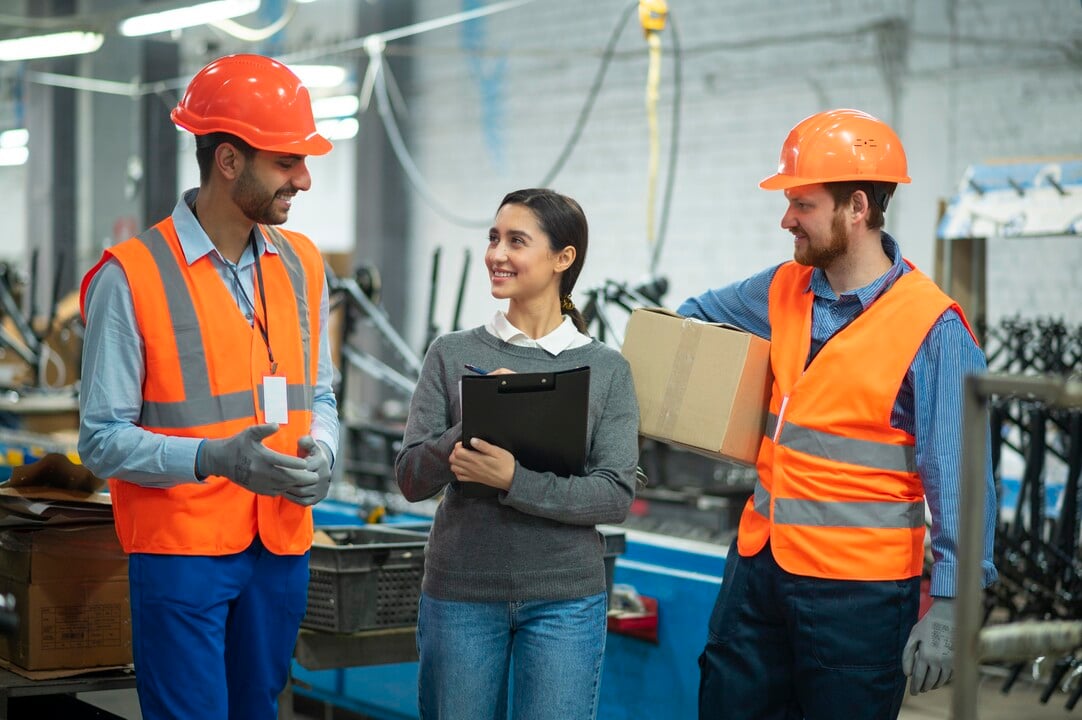 Three workers stand together in a factory. Two men wear orange safety vests, hard hats, and gloves; one holds a box. A woman in a gray sweater holds a clipboard and smiles as they talk. Industrial equipment is in the background.