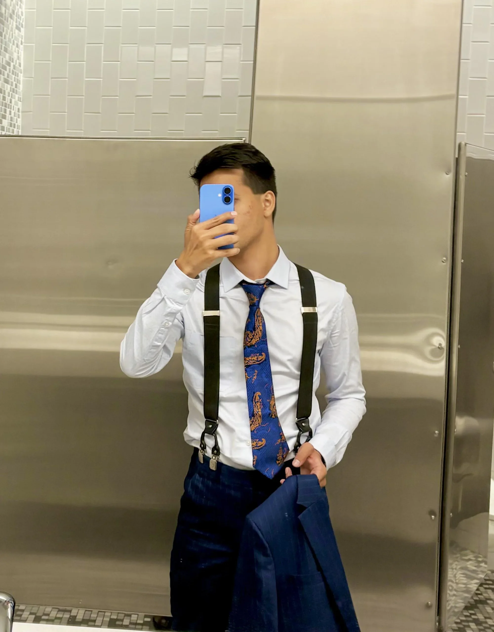 A person in a restroom takes a mirror selfie while wearing a white shirt, patterned tie, suspenders, and dark pants, holding a blue phone in front of their face and a blue jacket in their hand.