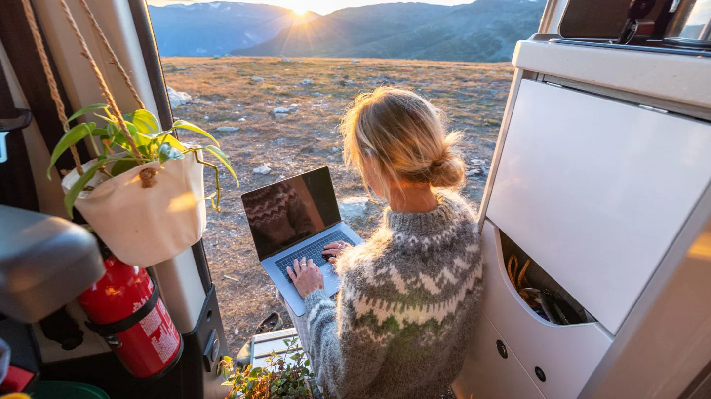 A person with blonde hair in a patterned sweater works on a laptop inside a camper van, facing the open door with a scenic mountain view and sunrise in the background. A plant and fire extinguisher are visible nearby.