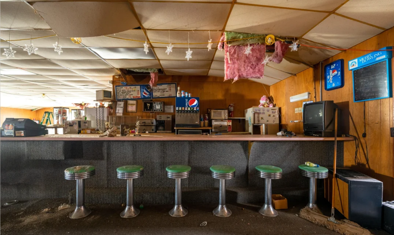 An abandoned diner with a row of six green-topped stools, a damaged ceiling, dusty counter, old menus, and a Pepsi soda fountain. The scene appears neglected, with ceiling tiles hanging down and pink insulation exposed.