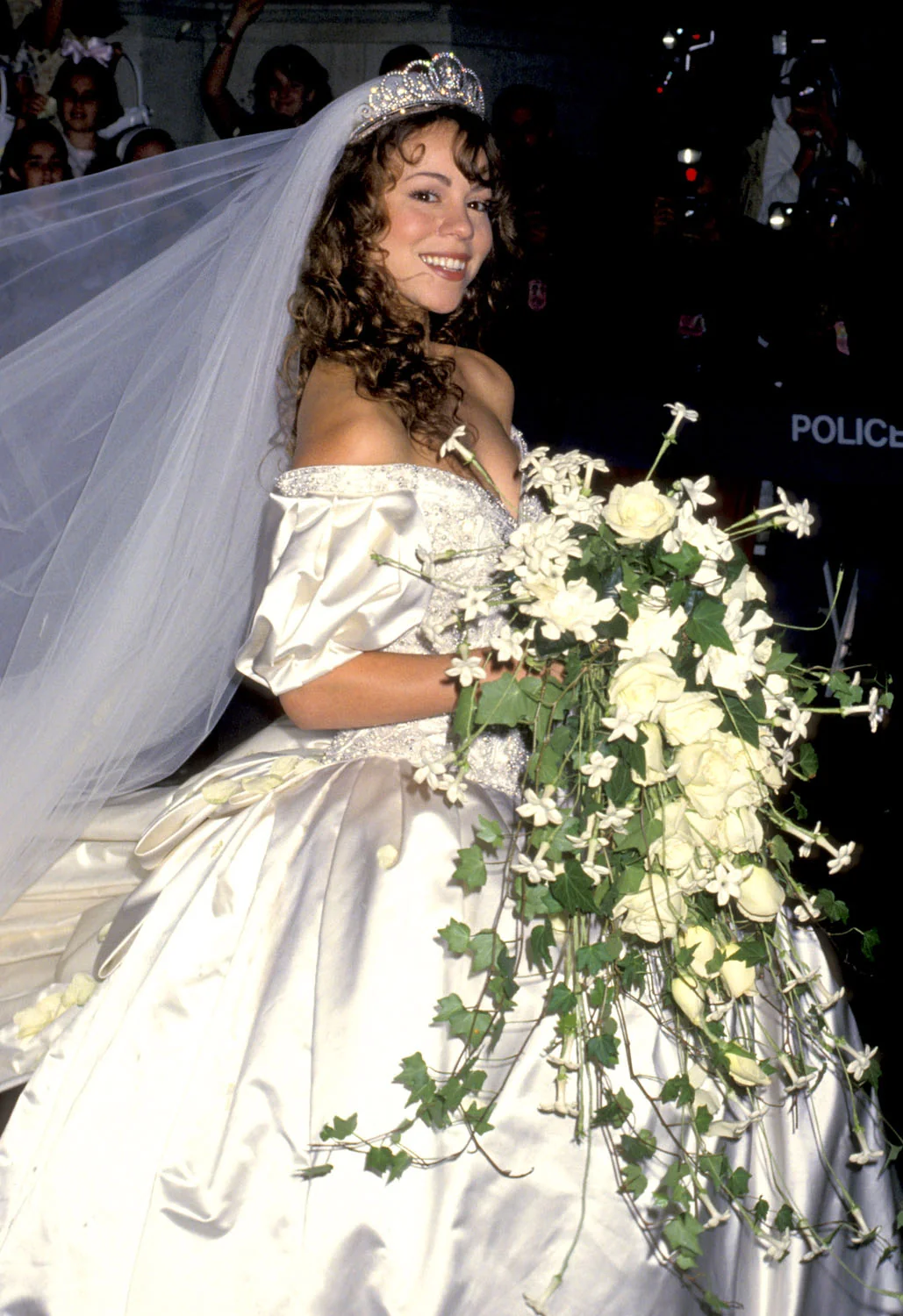 A woman in an ornate white wedding dress and tiara smiles, holding a large cascading bouquet of white flowers and greenery. She stands outdoors, surrounded by people and photographers.