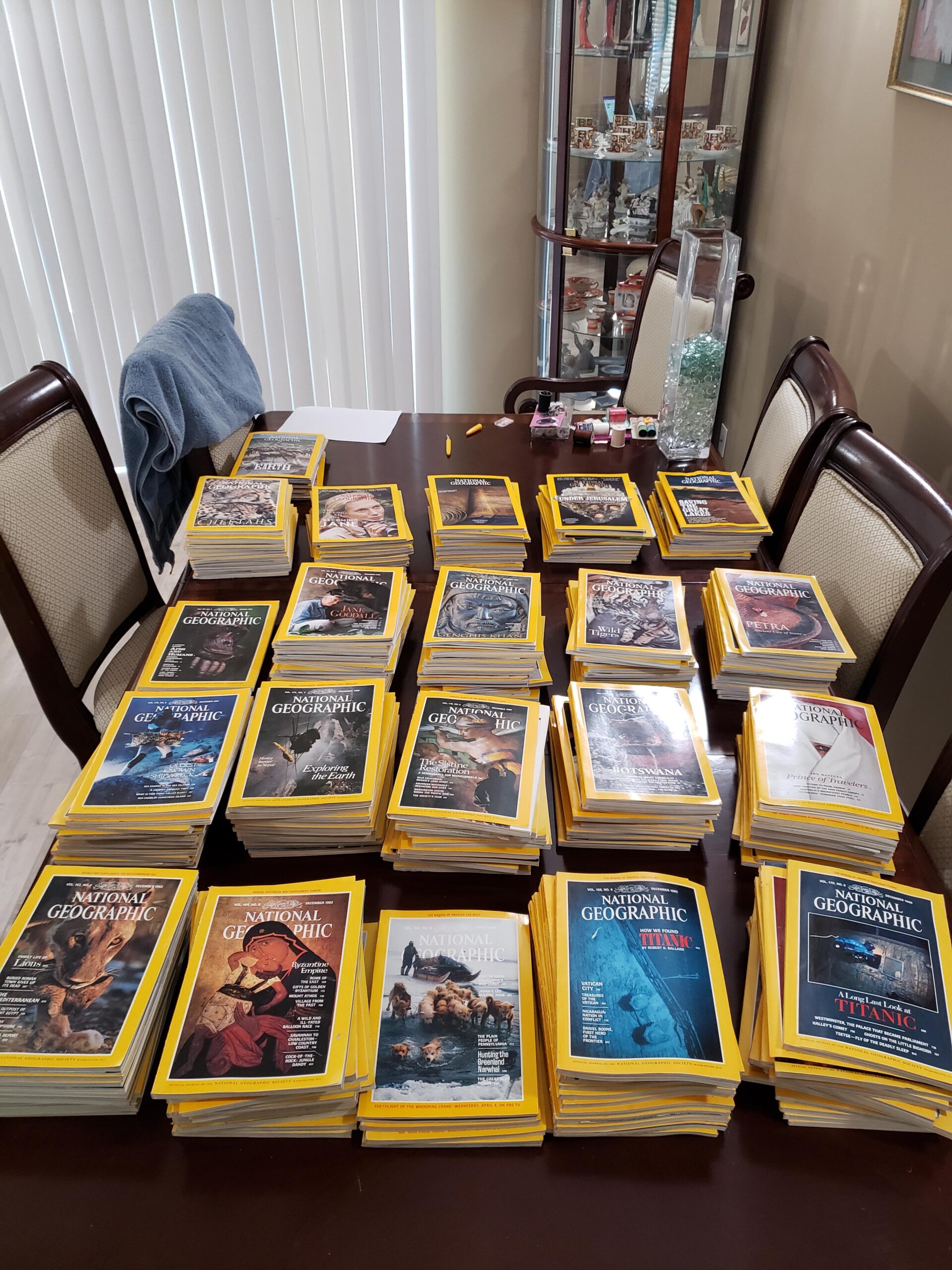 A dining table covered with neatly stacked National Geographic magazines, each displaying distinctive yellow borders and various cover images; chairs and a glass cabinet are visible in the background.