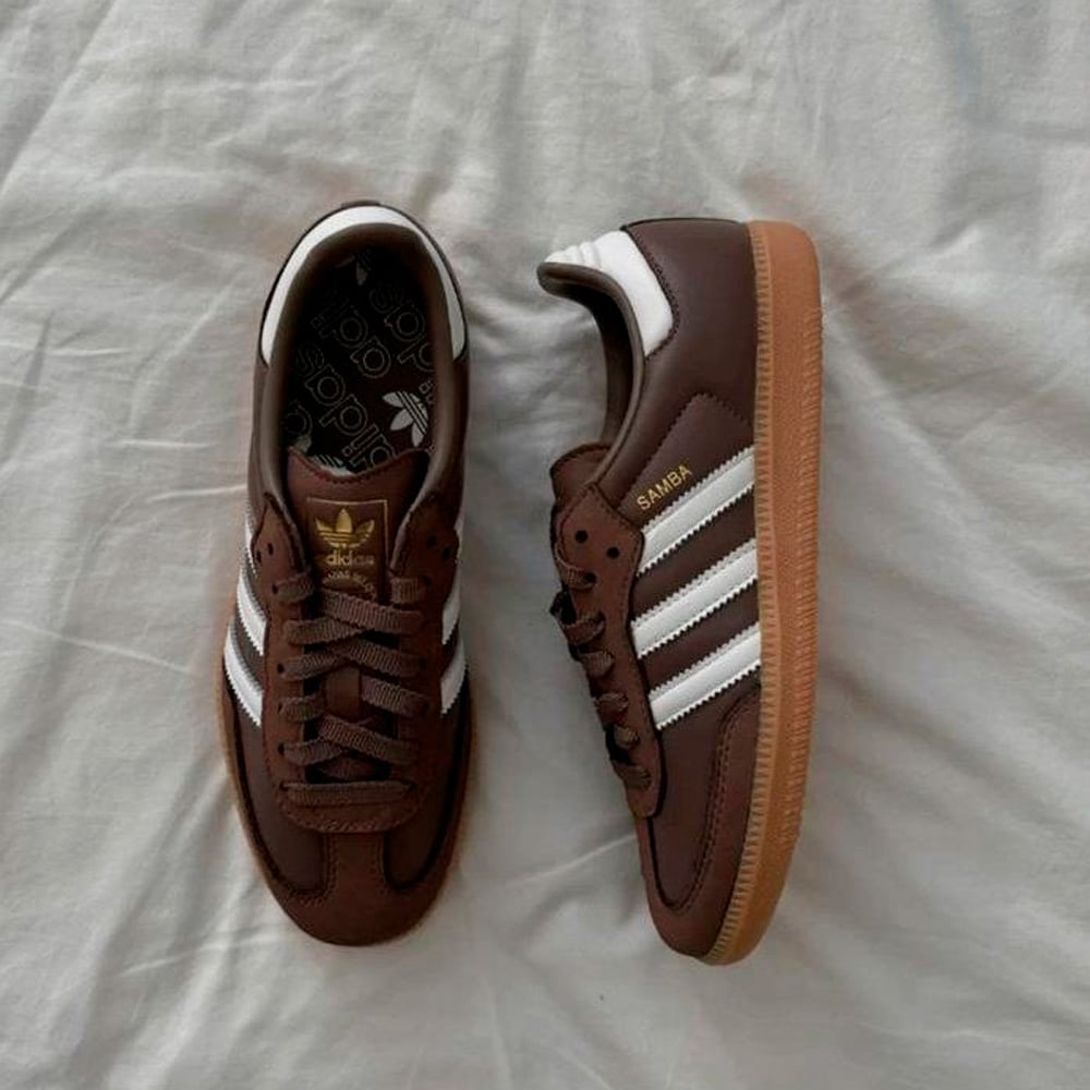A pair of brown Adidas Samba sneakers with white stripes and gold "SAMBA" lettering, placed on a white fabric background. One shoe is upright, the other is on its side.