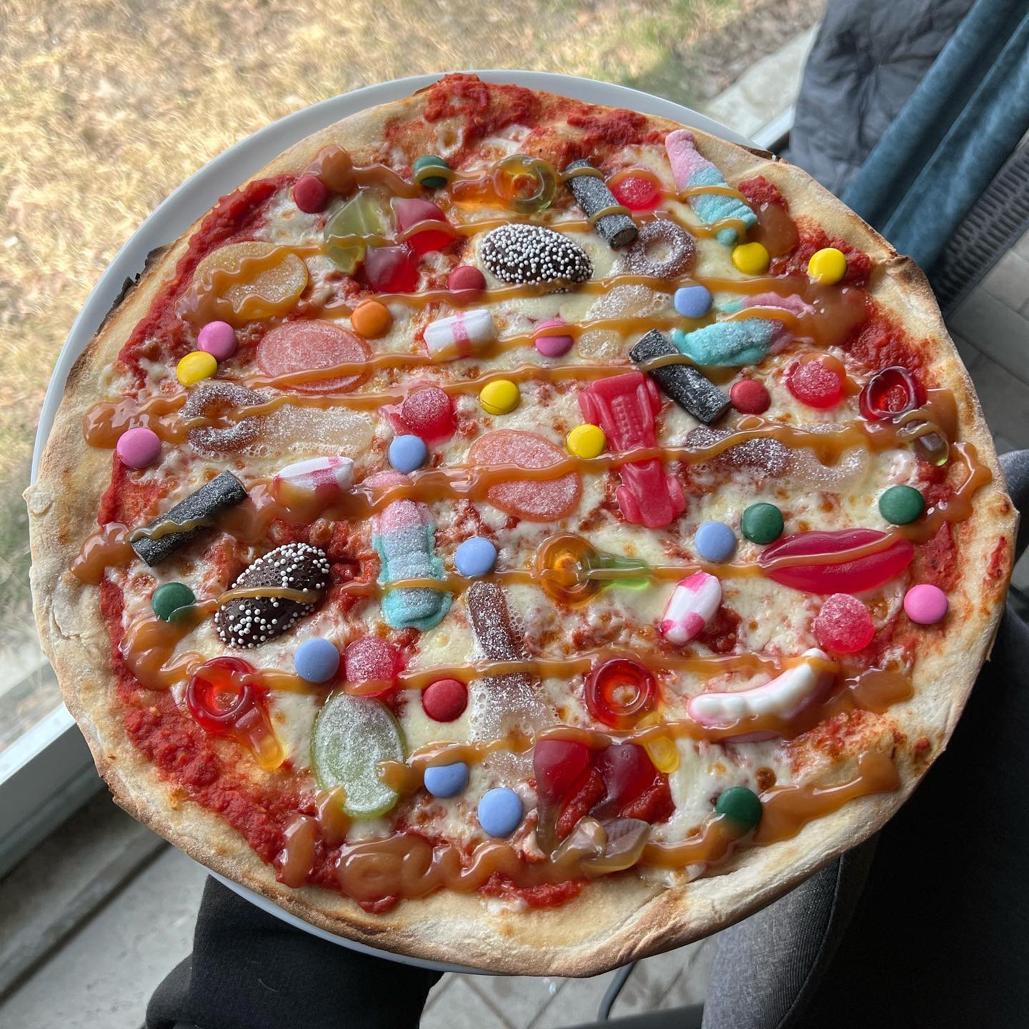 A pizza topped with tomato sauce, cheese, colorful gummy candies, chocolates, and caramel drizzle, held by someone near a window with grass visible outside.