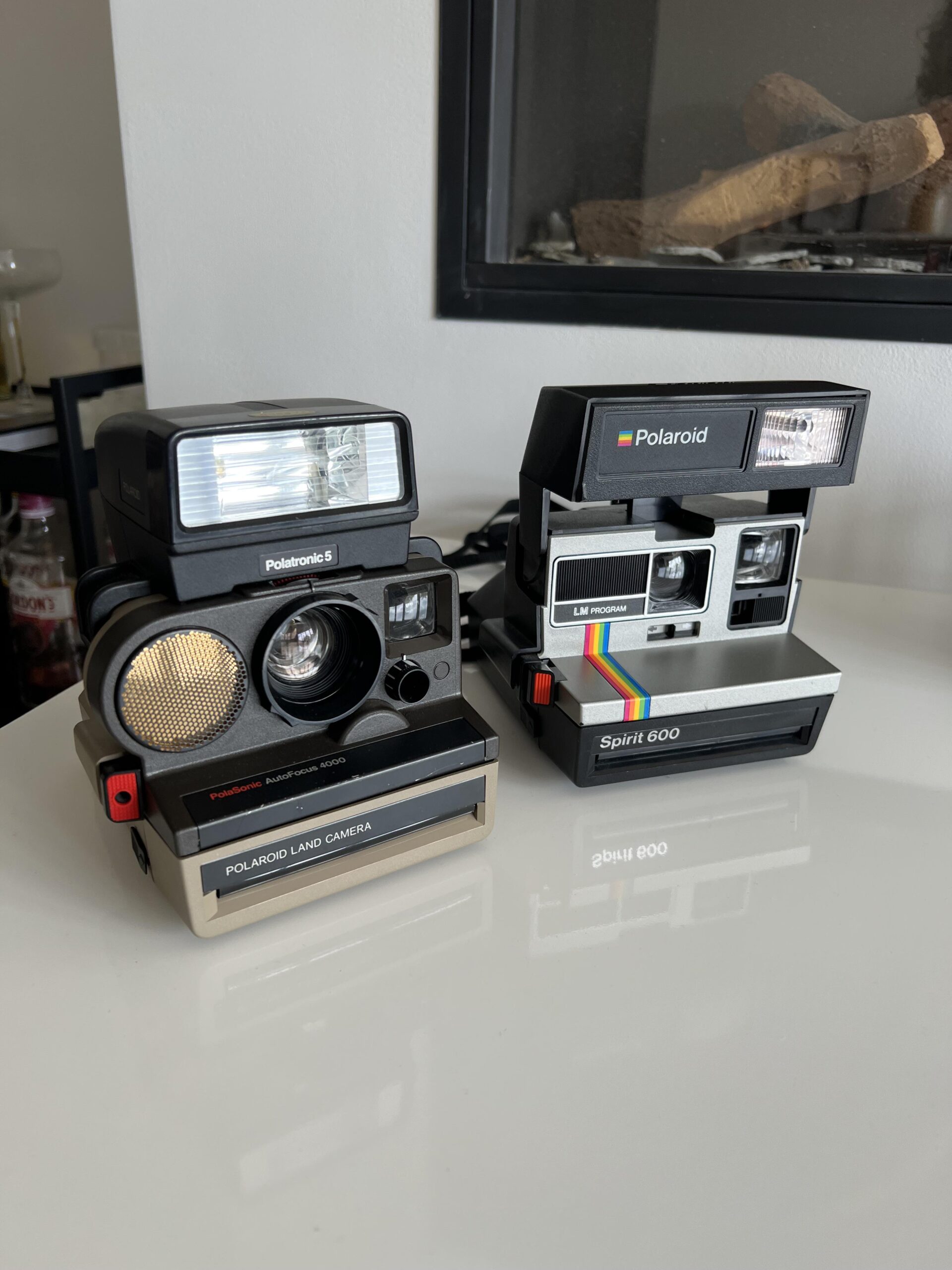 Two vintage Polaroid instant cameras sit side by side on a white surface; one is a Polaroid Land Camera, and the other is a Polaroid Spirit 600 with a rainbow stripe. Logs are visible in the background.