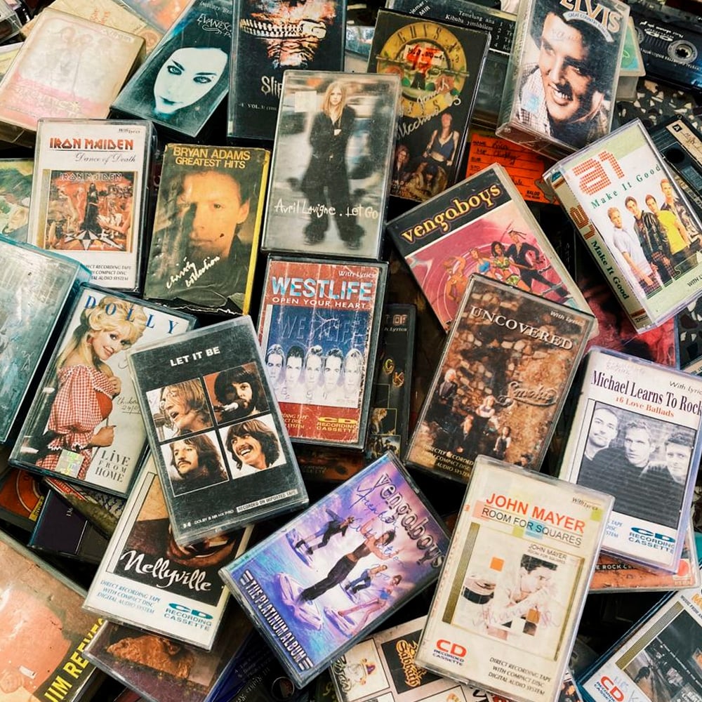 A scattered pile of old music cassette tapes and CD covers featuring various artists and bands, including Michael Jackson, Elvis Presley, Iron Maiden, Westlife, Vengaboys, and John Mayer.