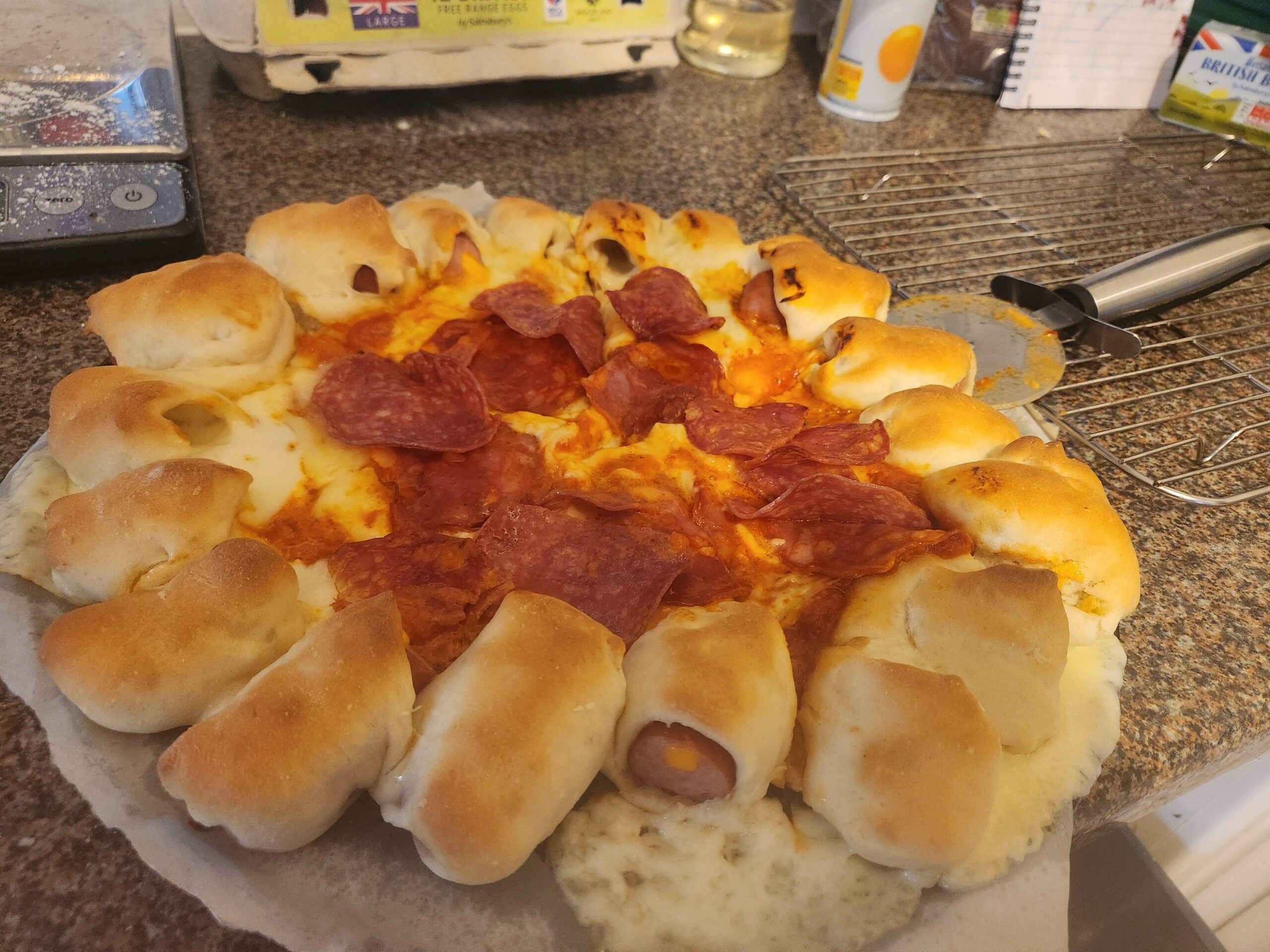 A homemade pizza with pepperoni slices in the center and mini sausage rolls baked into the crust, sitting on parchment paper on a kitchen counter. Cooking utensils and ingredients are visible in the background.