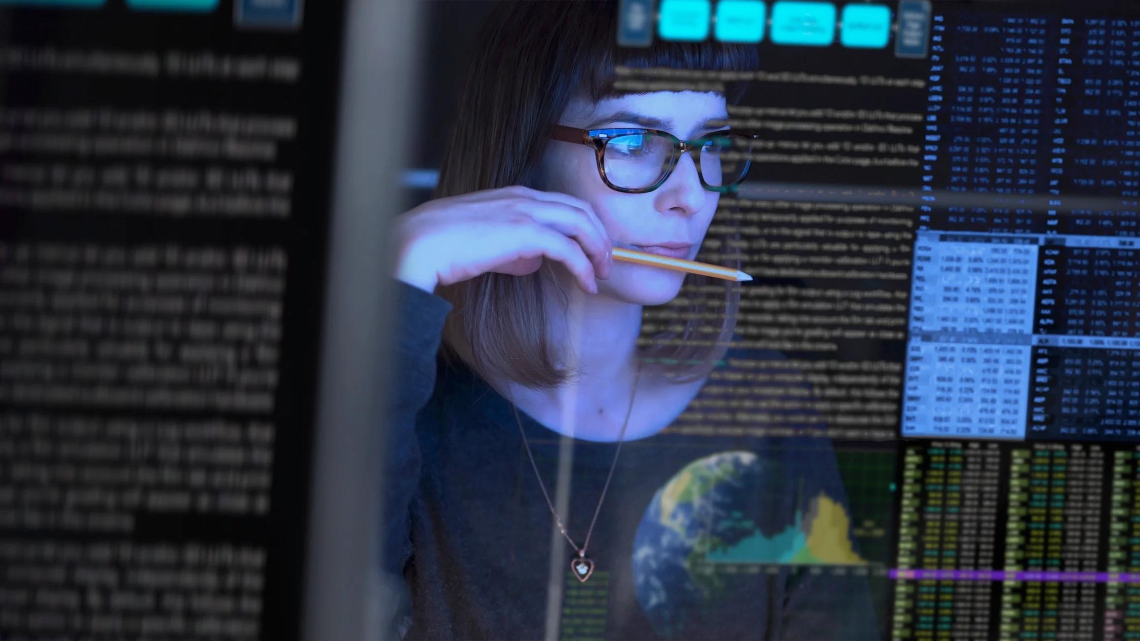A woman wearing glasses looks thoughtfully at computer screens displaying code, data, and graphs, holding a pencil to her lips. Reflections of digital information are visible on the glass in front of her.