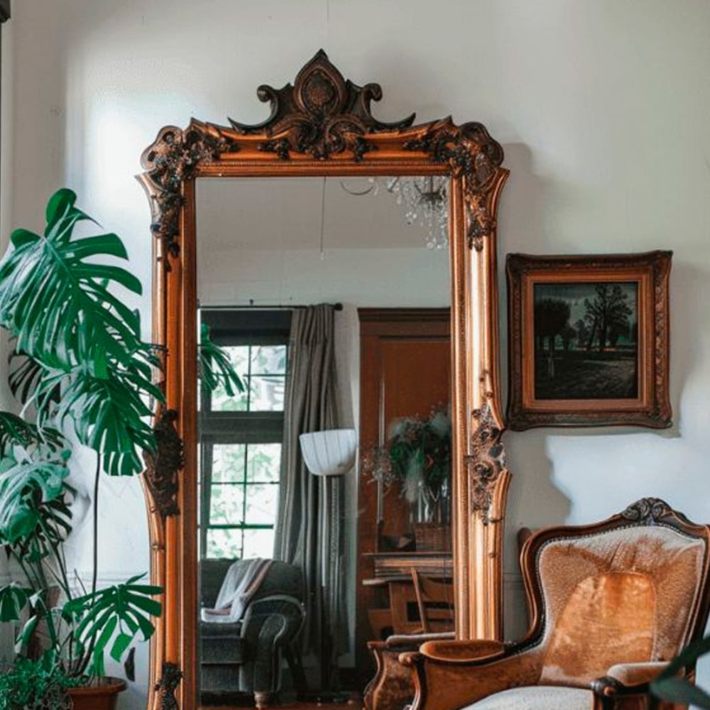 An ornate, gold-framed mirror stands against a white wall, reflecting a cozy living room with large windows, curtains, a chair, and plants. A leafy green plant and a framed picture hang nearby.
