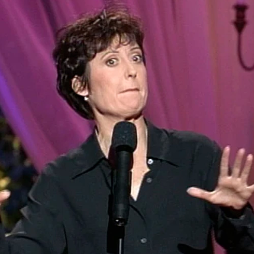 A person with short, curly dark hair stands on stage in front of a microphone, wearing a black shirt and gesturing with both hands. Pink curtains and a wall sconce are visible in the background.