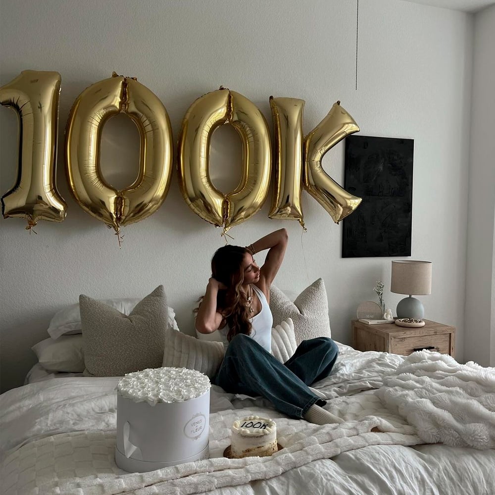 A woman sits on a bed with white bedding, stretching and smiling. Behind her are large gold balloons spelling "100K". On the bed are a white flower box and a small cake with "100K" written on it.